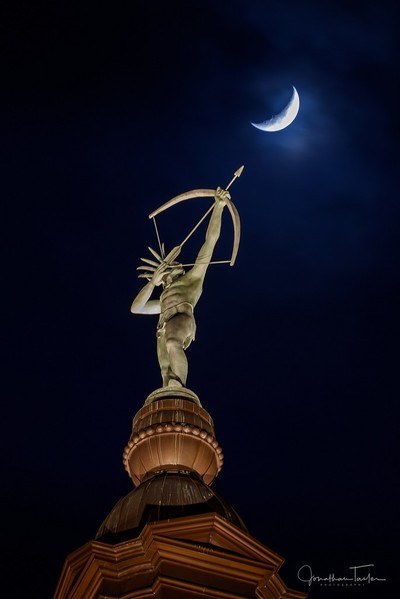 Ad Astra figure on the capitol dome of Topeka, Kansas aims a drawn bow toward a crescent moon in the night sky. The moon is surrounded by a lighter blue than the night sky while the Ad Astra figure and the cupola it sits on are lit from below.