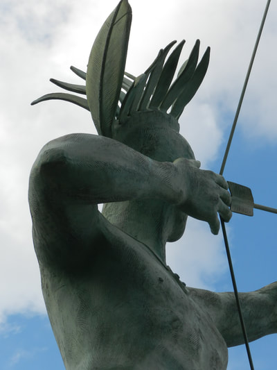 Close-up of the left back of the Ad Astra statue in McPherson, Kansas. The figure draws a bowstring back, hand blocking face. A crest of feathers tops his head.
